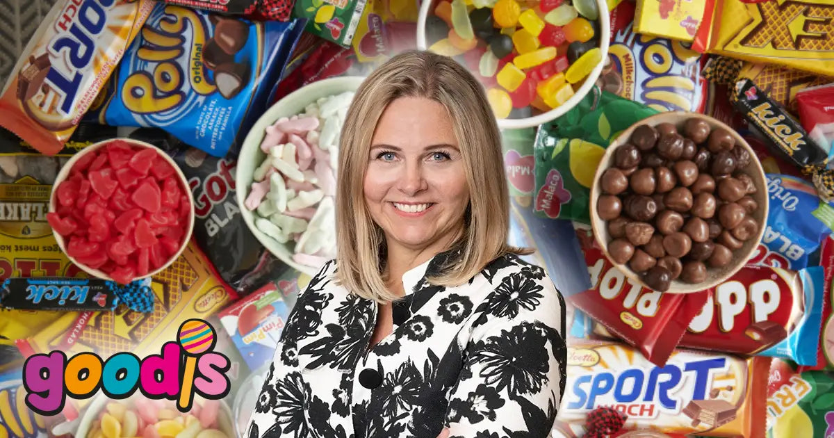 Woman smiling and looking into a camera with candy and chocolate behind her and the Goodis logo at the bottom left.